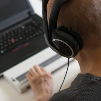 Person wearing large headphones using a laptop with a braille display, viewed from behind.
